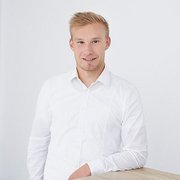 Professionelles Porträt eines lächelnden Mannes Ein junger Mann mit blonden Haaren lächelt freundlich, während er an einem hellen Tisch steht und in die Kamera schaut.