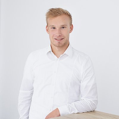 Ein junger Mann mit blonden Haaren lächelt freundlich, während er an einem hellen Tisch steht und in die Kamera schaut.